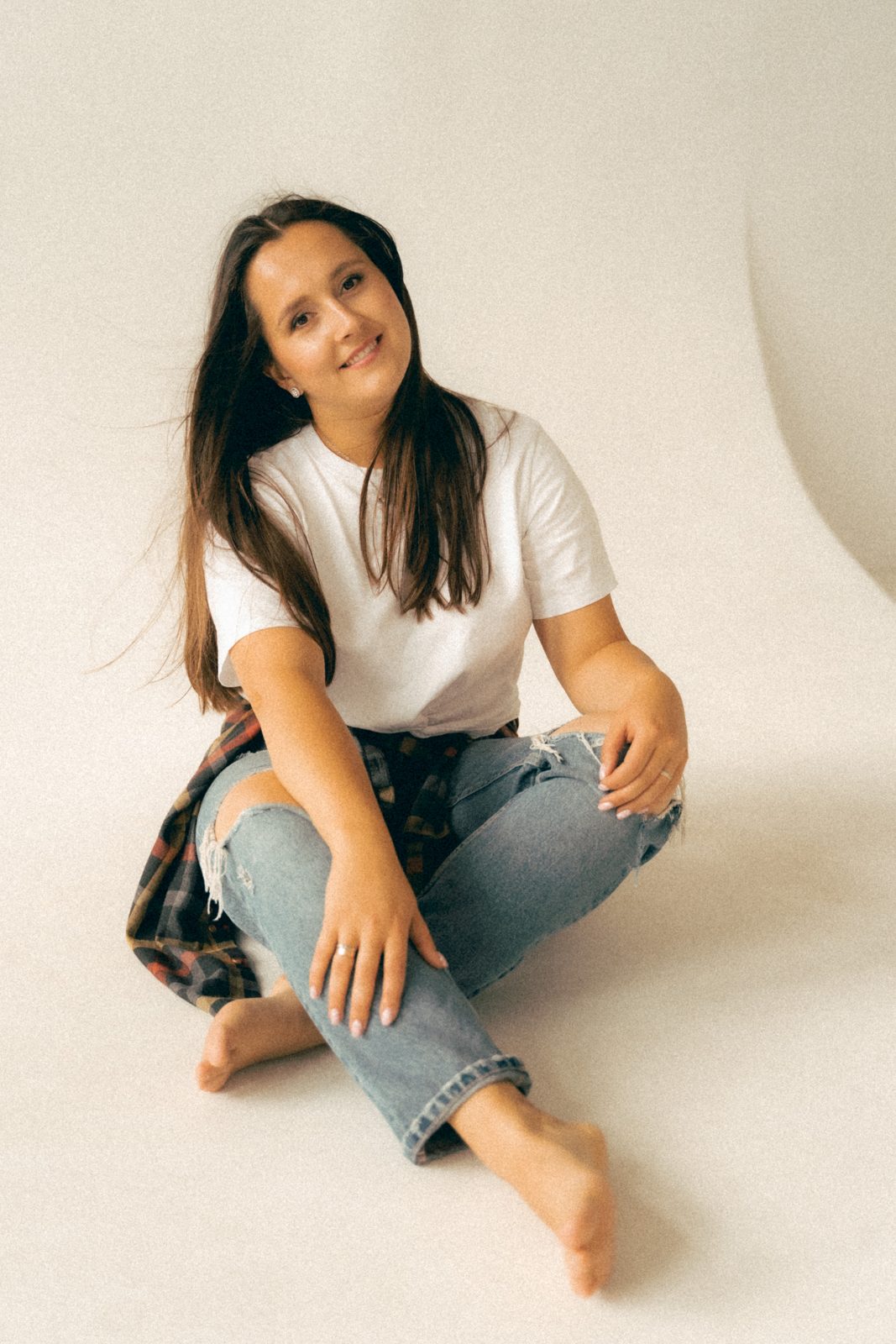 Woman in a white tee and jeans sitting barefoot on a cream studio backdrop