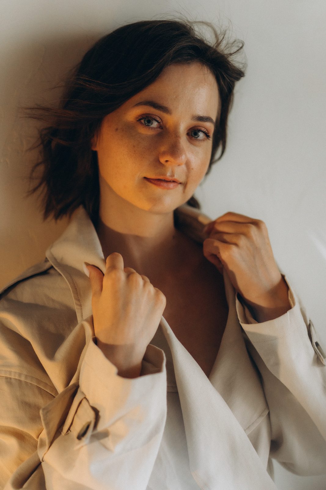 Woman with freckles holding the collar of a cream trench coat in warm light